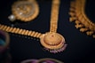 An intricate gold jewelry piece featuring a beautifully detailed circular pendant with red and purple gemstones set in ornate patterns. The surrounding elements include additional gold chains and embellishments, showcasing exquisite craftsmanship against a dark background.