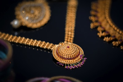 An intricate gold jewelry piece featuring a beautifully detailed circular pendant with red and purple gemstones set in ornate patterns. The surrounding elements include additional gold chains and embellishments, showcasing exquisite craftsmanship against a dark background.