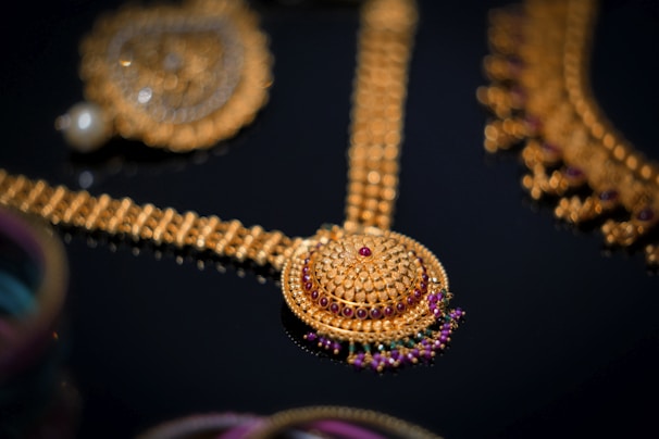 An intricate gold jewelry piece featuring a beautifully detailed circular pendant with red and purple gemstones set in ornate patterns. The surrounding elements include additional gold chains and embellishments, showcasing exquisite craftsmanship against a dark background.