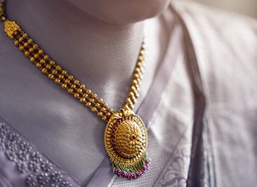 A close-up view of an ornate gold necklace with intricate designs and colorful gemstones, worn by a person dressed in a traditional garment with intricate patterns.