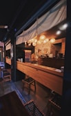 A cozy, dimly lit bar with wooden furniture and traditional Japanese decor. Lanterns hang from the ceiling, casting a warm glow over the long wooden counter. The walls feature wall art with Asian-inspired designs.