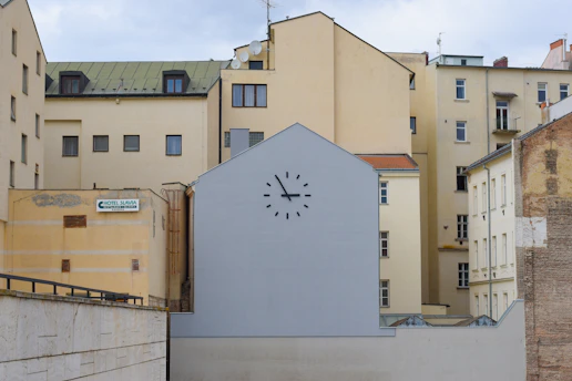 A group of beige and cream-colored apartment buildings with varied architectural styles. In the foreground, a flat building or wall painted grey displays a large clock without visible hands, positioned above small windows. On the left side, a sign reads 'Hotel Slavia', mounted on a yellow wall of the adjacent building.