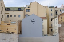 A group of beige and cream-colored apartment buildings with varied architectural styles. In the foreground, a flat building or wall painted grey displays a large clock without visible hands, positioned above small windows. On the left side, a sign reads 'Hotel Slavia', mounted on a yellow wall of the adjacent building.