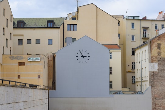 A group of beige and cream-colored apartment buildings with varied architectural styles. In the foreground, a flat building or wall painted grey displays a large clock without visible hands, positioned above small windows. On the left side, a sign reads 'Hotel Slavia', mounted on a yellow wall of the adjacent building.