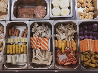 A variety of food items presented in stainless steel trays, featuring different colors and shapes. There are slices of processed meats, boiled and fried eggs, and various types of sausages. Some trays contain dim sum, colorful pastries, and sausage rolls. The arrangement is neat, suggesting a buffet or food service setting.