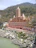 A large, multi-story temple with intricate architecture stands on the banks of a river, surrounded by lush green hills. The temple features numerous tiers with spires and is painted in shades of pink and orange. In the foreground, there are rocky riverbanks, and the water appears to be flowing gently.
