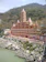A large, multi-story temple with intricate architecture stands on the banks of a river, surrounded by lush green hills. The temple features numerous tiers with spires and is painted in shades of pink and orange. In the foreground, there are rocky riverbanks, and the water appears to be flowing gently.