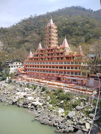 A large, multi-story temple with intricate architecture stands on the banks of a river, surrounded by lush green hills. The temple features numerous tiers with spires and is painted in shades of pink and orange. In the foreground, there are rocky riverbanks, and the water appears to be flowing gently.
