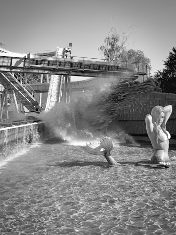 A large mermaid statue rises from the water in front of a log flume ride. The background features a dramatic splash created by logs descending the ride. The setting is surrounded by trees and other structures, with the log flume elevated above the water.