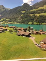 A picturesque village is nestled among lush green fields and surrounded by dramatic mountains under a clear blue sky. The tranquil turquoise lake reflects the natural beauty of the countryside. Charming wooden houses with sloped roofs are scattered throughout the landscape, and winding paths connect them, adding to the serene rural setting.