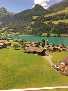 A picturesque village is nestled among lush green fields and surrounded by dramatic mountains under a clear blue sky. The tranquil turquoise lake reflects the natural beauty of the countryside. Charming wooden houses with sloped roofs are scattered throughout the landscape, and winding paths connect them, adding to the serene rural setting.