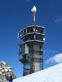 A large communication tower stands on a snowy mountain slope, featuring numerous antennas and satellite dishes. A person in a blue jacket is visible at the base, accentuating the size of the structure. The sky is clear and vibrant blue.