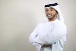 A man in traditional Middle Eastern attire is standing with his arms crossed, smiling confidently. He is wearing a white kandura and a ghutra with an agal.