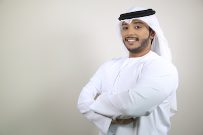 Stylish man wearing a black and gold thobe with intricate embroidery, smiling confidently in a minimalistic setting.