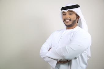 A man in traditional Middle Eastern attire is standing with his arms crossed, smiling confidently. He is wearing a white kandura and a ghutra with an agal.