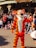 A person dressed in a bright orange and black striped costume resembling a tiger character is interacting with a crowd of people. The scene is lively, with many adults and children sitting or standing, taking pictures and watching. The background features a charming building with a sign that reads 'Unique Flora's Boutique'.