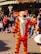 A person dressed in a bright orange and black striped costume resembling a tiger character is interacting with a crowd of people. The scene is lively, with many adults and children sitting or standing, taking pictures and watching. The background features a charming building with a sign that reads 'Unique Flora's Boutique'.