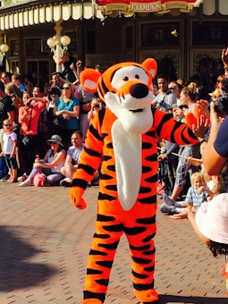 A person dressed in a bright orange and black striped costume resembling a tiger character is interacting with a crowd of people. The scene is lively, with many adults and children sitting or standing, taking pictures and watching. The background features a charming building with a sign that reads 'Unique Flora's Boutique'.