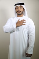 A man is wearing traditional Middle Eastern attire, including a white kandura and a keffiyeh. His right hand is placed over his heart, and he is standing against a plain, light-colored background.