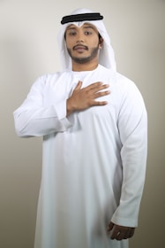 A man is wearing traditional Middle Eastern attire, including a white kandura and a keffiyeh. His right hand is placed over his heart, and he is standing against a plain, light-colored background.