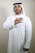 A man is wearing traditional Middle Eastern attire, including a white kandura and a keffiyeh. His right hand is placed over his heart, and he is standing against a plain, light-colored background.