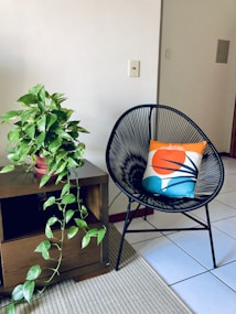 A cozy room corner features a black wireframe chair with a colorful cushion placed on it, displaying a design with orange, blue, and white colors. Beside the chair is a wooden side table, on which a leafy green potted plant rests. The plant's vines cascade over the side of the table. The floor is covered with white tiles and a beige woven rug, contributing to a minimalist and natural aesthetic.