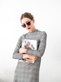 A person wearing sunglasses and a plaid dress holds a fashion magazine featuring a cover with a woman in a stylish outfit. The background is plain, enhancing the minimalist and modern aesthetic.