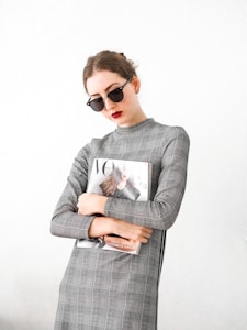 A person wearing sunglasses and a plaid dress holds a fashion magazine featuring a cover with a woman in a stylish outfit. The background is plain, enhancing the minimalist and modern aesthetic.