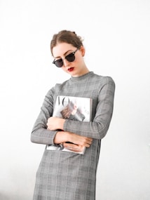 A person wearing sunglasses and a plaid dress holds a fashion magazine featuring a cover with a woman in a stylish outfit. The background is plain, enhancing the minimalist and modern aesthetic.