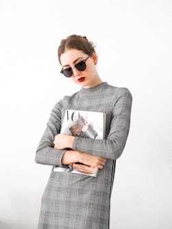 A person wearing sunglasses and a plaid dress holds a fashion magazine featuring a cover with a woman in a stylish outfit. The background is plain, enhancing the minimalist and modern aesthetic.