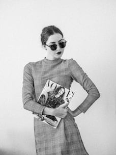 A woman wearing a checkered dress and dark sunglasses holds a fashion magazine, displaying a confident and stylish demeanor. The image is in black and white, emphasizing contrasts.