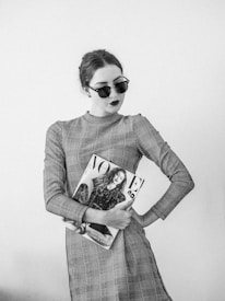A woman wearing a checkered dress and dark sunglasses holds a fashion magazine, displaying a confident and stylish demeanor. The image is in black and white, emphasizing contrasts.