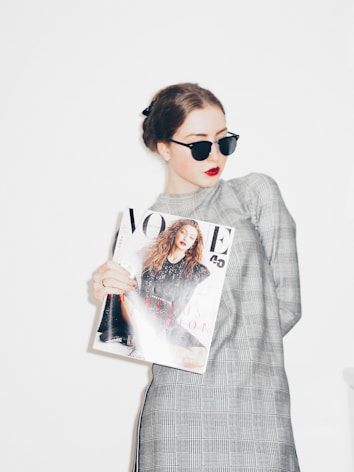 A person wearing sunglasses and red lipstick holds a fashion magazine while dressed in a checkered long-sleeve outfit. Their hair is styled into a neat bun.