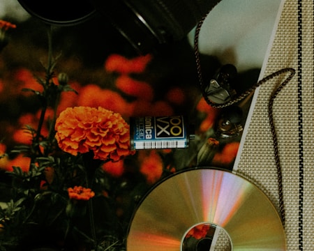 A collection of objects displayed on a patterned surface includes a vibrant orange marigold, a film roll with blue and white labeling, a CD that reflects light in rainbow colors, and a pair of in-ear headphones with a braided cord. The background is filled with blurred orange flowers.
