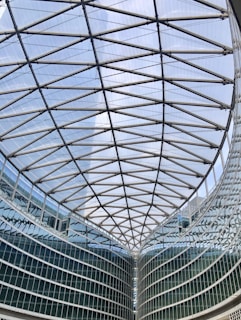 A large modern glass building with a geometric, lattice-like ceiling structure. The architecture features steel beams forming an intricate pattern above a curved glass facade.