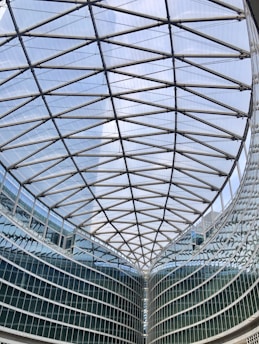 A large modern glass building with a geometric, lattice-like ceiling structure. The architecture features steel beams forming an intricate pattern above a curved glass facade.
