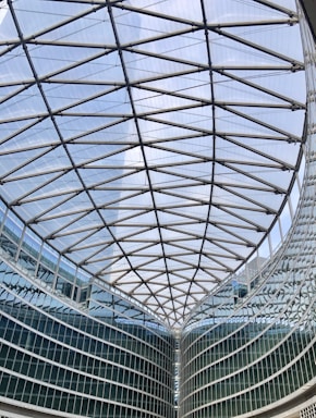 A large modern glass building with a geometric, lattice-like ceiling structure. The architecture features steel beams forming an intricate pattern above a curved glass facade.