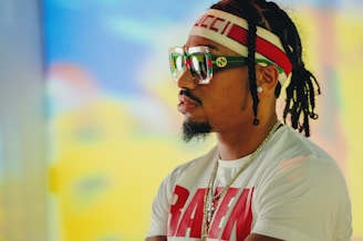A person with braided hair wearing a white t-shirt with the word RAVEN, a headband, and oversized sunglasses. The headband and sunglasses feature a designer logo. The background is blurred with vibrant colors.