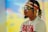 A person with braided hair wearing a white t-shirt with the word RAVEN, a headband, and oversized sunglasses. The headband and sunglasses feature a designer logo. The background is blurred with vibrant colors.