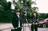 Two people stand next to a professional video camera mounted on a tripod, with one person dressed in a black jacket and cap, while the other wears a colorful hoodie. They appear to be in an outdoor setting with greenery as the backdrop, and a red car is parked nearby.