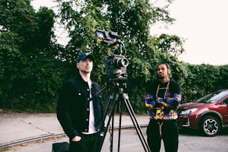 Two people stand next to a professional video camera mounted on a tripod, with one person dressed in a black jacket and cap, while the other wears a colorful hoodie. They appear to be in an outdoor setting with greenery as the backdrop, and a red car is parked nearby.