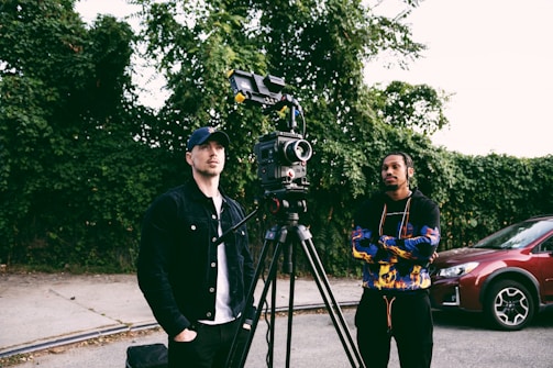 Two people stand next to a professional video camera mounted on a tripod, with one person dressed in a black jacket and cap, while the other wears a colorful hoodie. They appear to be in an outdoor setting with greenery as the backdrop, and a red car is parked nearby.