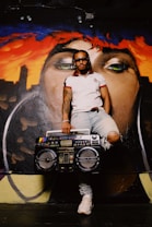 A person with sunglasses and braids is standing against a vibrant mural depicting a close-up of a face. Their outfit includes a white polo shirt and ripped jeans, and they hold a large retro boom box.