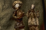 Two skeletal figurines are adorned in traditional attire made of textured material. One figure holds a pot, and both are positioned against a fabric-like backdrop, giving an antiquated and eerie aesthetic.