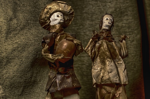 Two skeletal figurines are adorned in traditional attire made of textured material. One figure holds a pot, and both are positioned against a fabric-like backdrop, giving an antiquated and eerie aesthetic.