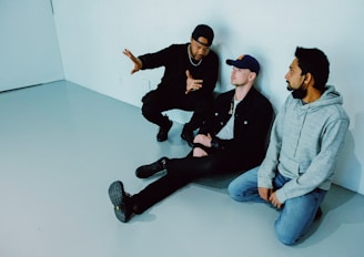 Three men are seated on the floor against a stark white wall. The man on the left wears a black outfit with a cap and gestures animatedly to the other two. The man in the middle, wearing a denim jacket and cap, listens attentively, while the man on the right in a gray hoodie sits quietly, looking toward the speaker.