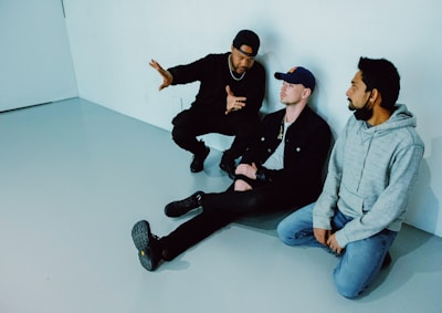 Three men are seated on the floor against a stark white wall. The man on the left wears a black outfit with a cap and gestures animatedly to the other two. The man in the middle, wearing a denim jacket and cap, listens attentively, while the man on the right in a gray hoodie sits quietly, looking toward the speaker.