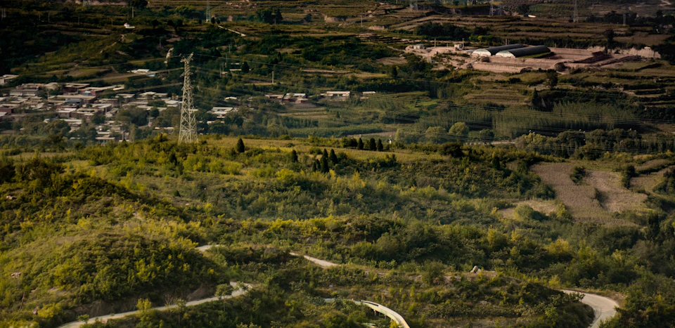 The image depicts a picturesque landscape with a winding road cutting through a lush, green valley. The road is surrounded by an abundance of trees and vegetation, creating a sense of tranquility and isolation. In the distance, there are several buildings scattered across the landscape, adding depth to the scene. The predominant colors in the image are shades of green from the vegetation and browns from the earthy tones of the valley floor and road.