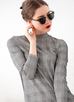 A person with dark hair is wearing sunglasses and a high-neck checkered top while posing thoughtfully. The background is plain, emphasizing the fashion elements and facial expression.