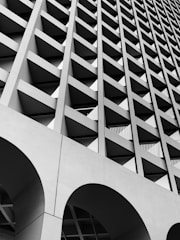 A striking architectural facade characterized by a geometric pattern of recessed square openings. The structure is composed of grid-like concrete beams, creating a sense of depth and rhythm. Arched sections can be seen at the bottom, contributing to the overall modernist aesthetic.